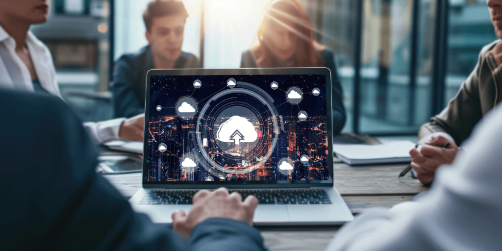Laptop displaying cloud technology icons on a table in a modern office. Four people in business attire sit in the background, engaged in discussion.