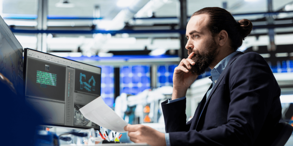 A man with a beard is focused on computer monitors displaying technical graphics, sitting in a modern office space, exuding concentration and professionalism.