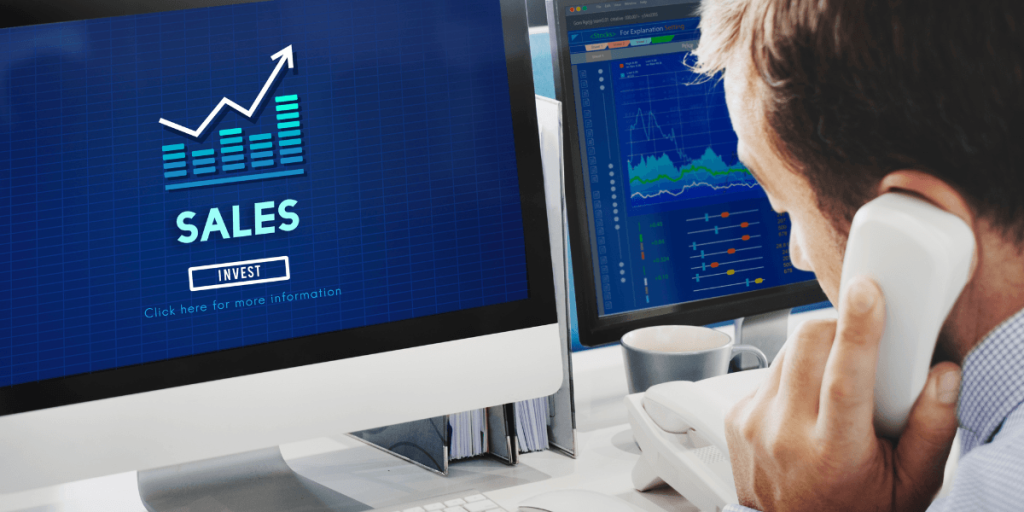A man on the phone in an office setting focuses on computer monitors. One displays a "Sales" graph trending upward; the other shows detailed charts.