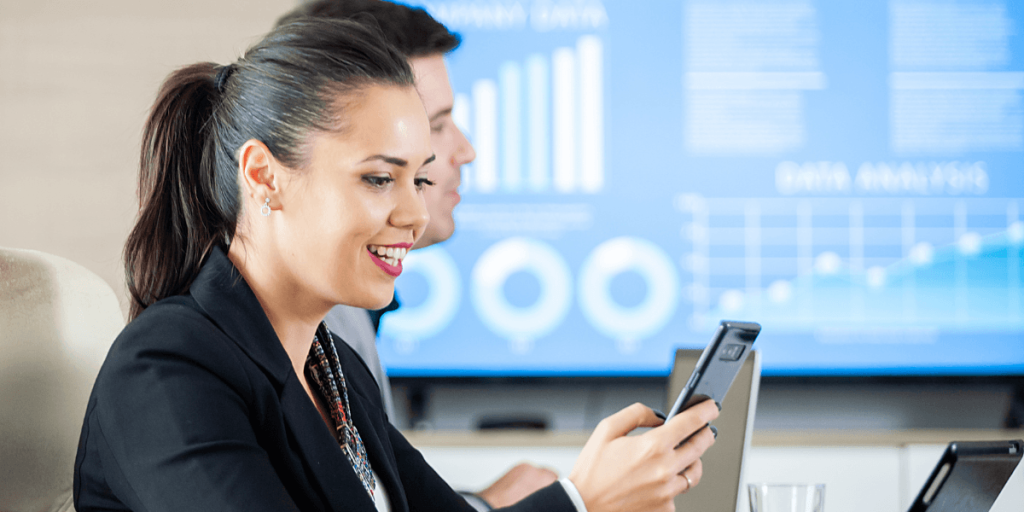 Smiling woman in a business suit uses a smartphone in an office, with blurred graphs on a screen in the background, suggesting data analysis.